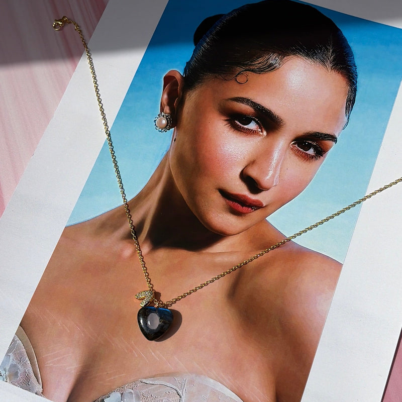 A gold chain necklace with a dark heart-shaped pendant is displayed on a printed fashion photograph of a woman wearing it, set against a pink draped fabric background.