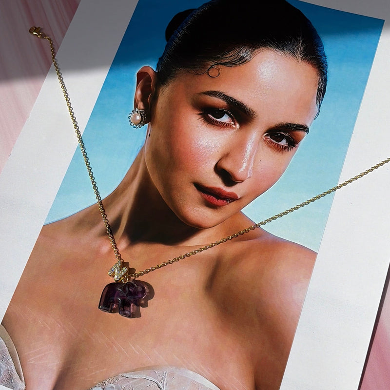 A gold chain necklace with a dark purple elephant pendant is displayed on a printed fashion photograph of a woman, set against a pink marbled background.